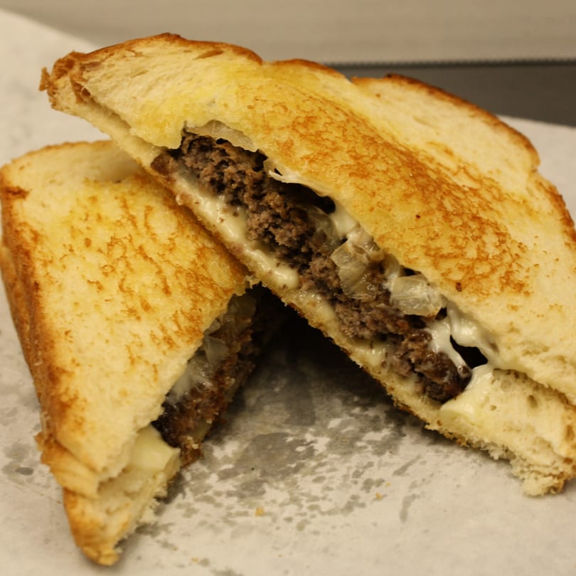 A Joe's To Go patty melt, cut in half, with melted cheese, grilled onions, and toasted bread.