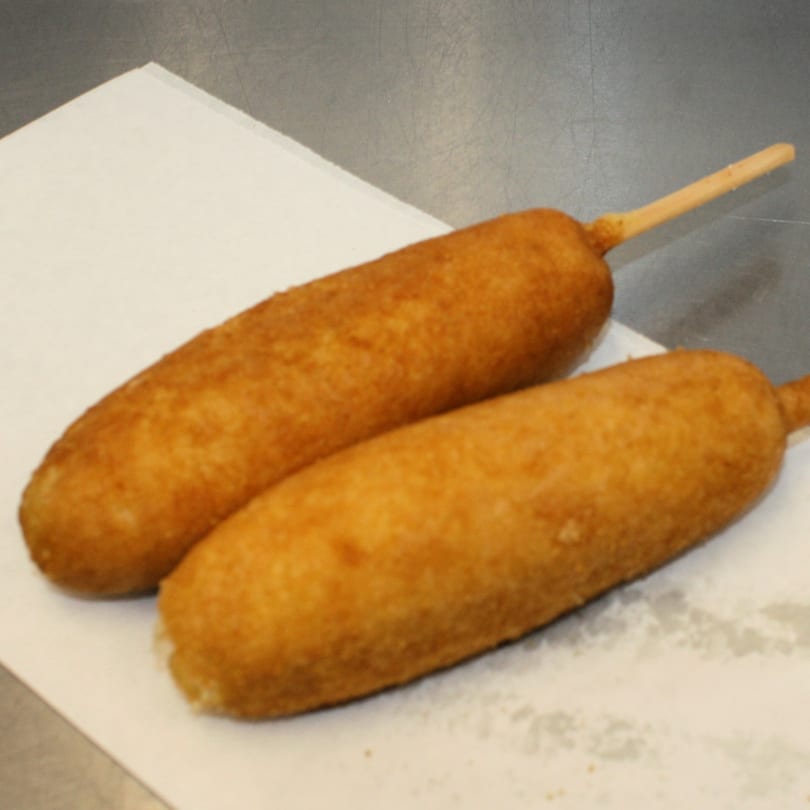 Two golden-brown corn dogs on sticks, resting on a white paper napkin at Joe's To Go.