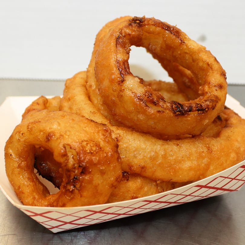 Golden-brown, crispy onion rings from Joe's To Go Drive Thru, served in a white paper tray.